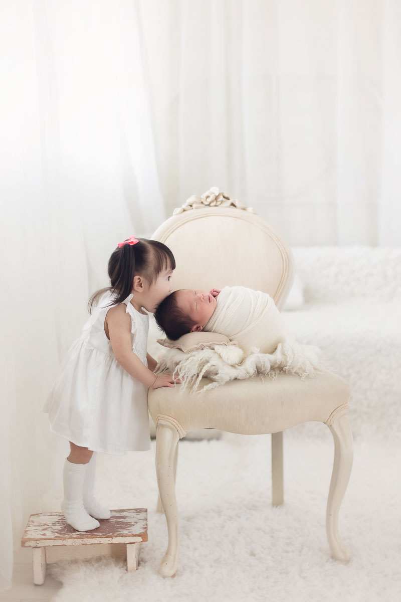 sister is kissing her new baby brother in a white and natural light studio